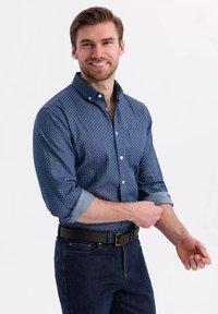 Navyblauw overhemd met knoopjes en kleine witte stippen, opgerolde mouwen en een borstzak, gedragen met donkere jeans en een zwarte riem.