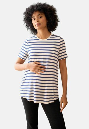 Frau mit lockigem Haar, die ein blau-weiß gestreiftes Hemd trägt, hält sanft ihren schwangeren Bauch und steht vor einem einfarbigen Hintergrund.