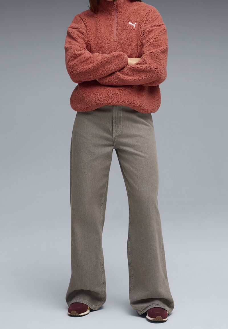 Personne portant un pull polaire couleur rouille avec un logo Puma blanc, un jean gris à jambes larges et des baskets bordeaux, debout les bras croisés.