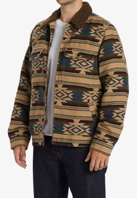 Tan patterned jacket featuring geometric designs in brown and teal, with a faux fur collar, two chest pockets, and a relaxed fit.
