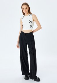 Top corto blanco sin mangas con acentos de lazo negro, combinado con pantalones de pierna ancha negros de talle alto. La modelo está de pie contra un fondo liso.