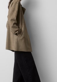 Mujer con una chaqueta de ante ligera y suelta de color marrón claro, con las manos en los bolsillos y pantalones negros, de pie contra un fondo gris claro liso.