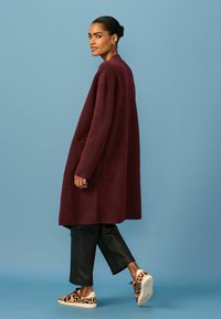 Bordeauxfarbener langer Mantel aus strukturiertem Stoff, mit seitlichen Taschen, getragen über schwarzen Lederhosen und Sneakers mit Leopardenmuster, vor einem blauen Hintergrund.