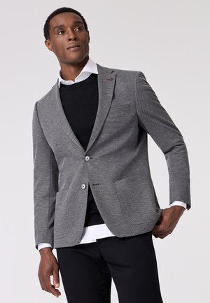 Grauer Blazer mit strukturiertem Finish, zwei Knöpfen und aufgesetzten Taschen. Getragen über einem schwarzen Pullover und einem weißen Hemd mit Kragen. Schwarze Hosen sichtbar.