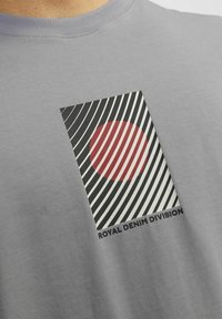 Grijze t-shirt met korte mouwen met een grafisch ontwerp van een rode cirkel, zwart-witte diagonale strepen en de tekst "ROYAL DENIM DIVISION."