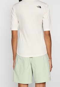 Krátké rukávy, krémově zbarvená košile se strukturovanou látkou a malým černým logem na zádech; doplněná světlými zelenými šortkami.