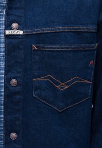 Temno modra jeans jakna z oranžnim šivanjem, žep spredaj s geometrijskim vzorcem šiva, kovinskimi gumbi in belo etiketo z napisom "REPLAY".