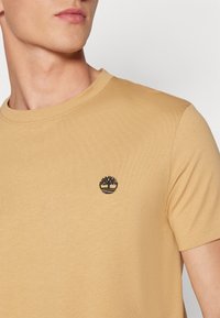 Kortærmet t-shirt med rund hals i lys tan-farvet bomuld, med et sort broderet trælogo på brystet. Blød tekstur, enkel design.