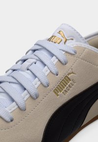 Κοντινό πλάνο μπεζ σουέντ αθλητικού παπουτσιού με ανοιχτό γκρι κορδόνια, μαύρη πλευρική ρίγα και χρυσό λογότυπο Puma Tackle στο πλάι και στη γλώσσα.