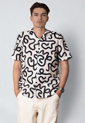 Mężczyzna w białej, krótkorękawowej koszuli z czarnymi abstrakcyjnymi falistymi wzorami oraz jasnobeżowych spodniach, z rękami w kieszeniach.
