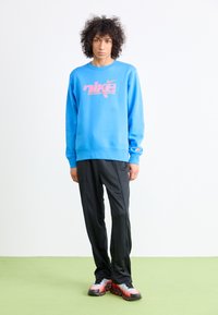 Blå bomulds sweatshirt med pink "nike" logo, sorte træningsbukser og multicolore sportssko på en grøn overflade. Simpelt baggrund.