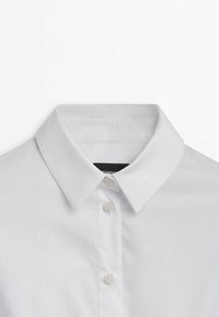 Chemise blanche à col pointu avec trois boutons visibles sur la patte de boutonnage devant, posée à plat sur un fond blanc.