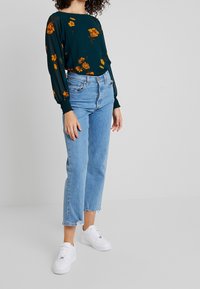 Blouse florale verte à manches longues, associée à un jean bleu clair taille haute et des baskets blanches. Détails subtils texturés sur les manches.