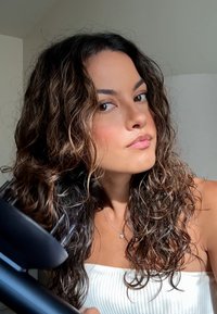 Secador de pelo negro sostenido al lado de una persona con cabello largo y rizado de color castaño. La persona lleva una parte de arriba sin tirantes blanca y tiene un fondo neutro.