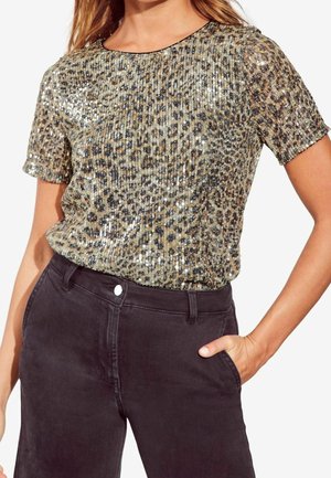 Kobieta w krótkim rękawie, złocisto-czarnym cekinowym topie w panterkę, włożonym do wysokich ciemnych spodni, z jedną ręką w kieszeni.