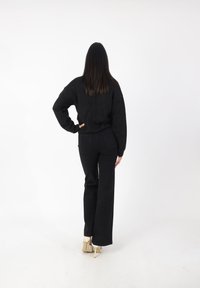 Svart oversize tröja med sänkta axlar, kombinerad med högmidjade svarta byxor. Modellen står med ryggen mot kameran, iförd beige klackar.