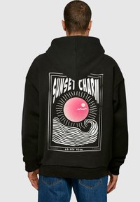 Felpa nera con cappuccio caratterizzata da una grande grafica sul retro con un design di sole rosa e onde, accentuata da testo e linee bianche, realizzata in tessuto morbido.
