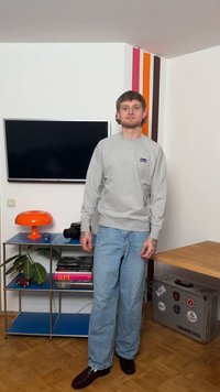 Giovane uomo in piedi in una stanza con una TV a parete, mensole blu che tengono libri, una lampada arancione, una macchina fotografica e una pianta.