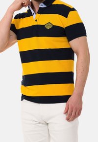 Homme portant un polo jaune et noir à rayures horizontales avec col et pantalon blanc, main touchant le col.