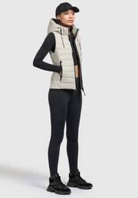 Hellgraue Steppweste mit Kapuze, ausgestattet mit Reißverschlusstaschen und geripptem Saum, getragen über einem schwarzen Langarmoberteil und schwarzen Leggings mit schwarzen Sneakers.
