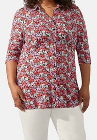 Camicia floreale bottone con sfondo nero e fiori rossi, rosa e bianchi, maniche a tre quarti e vestibilità leggermente ampia.