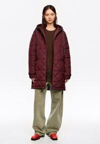 Bordowy pikowany długi płaszcz z kapturem, szare sznurki, w połączeniu z brązową bluzką i szerokimi zielonymi spodniami, uzupełniony czerwonymi butami w wzory.