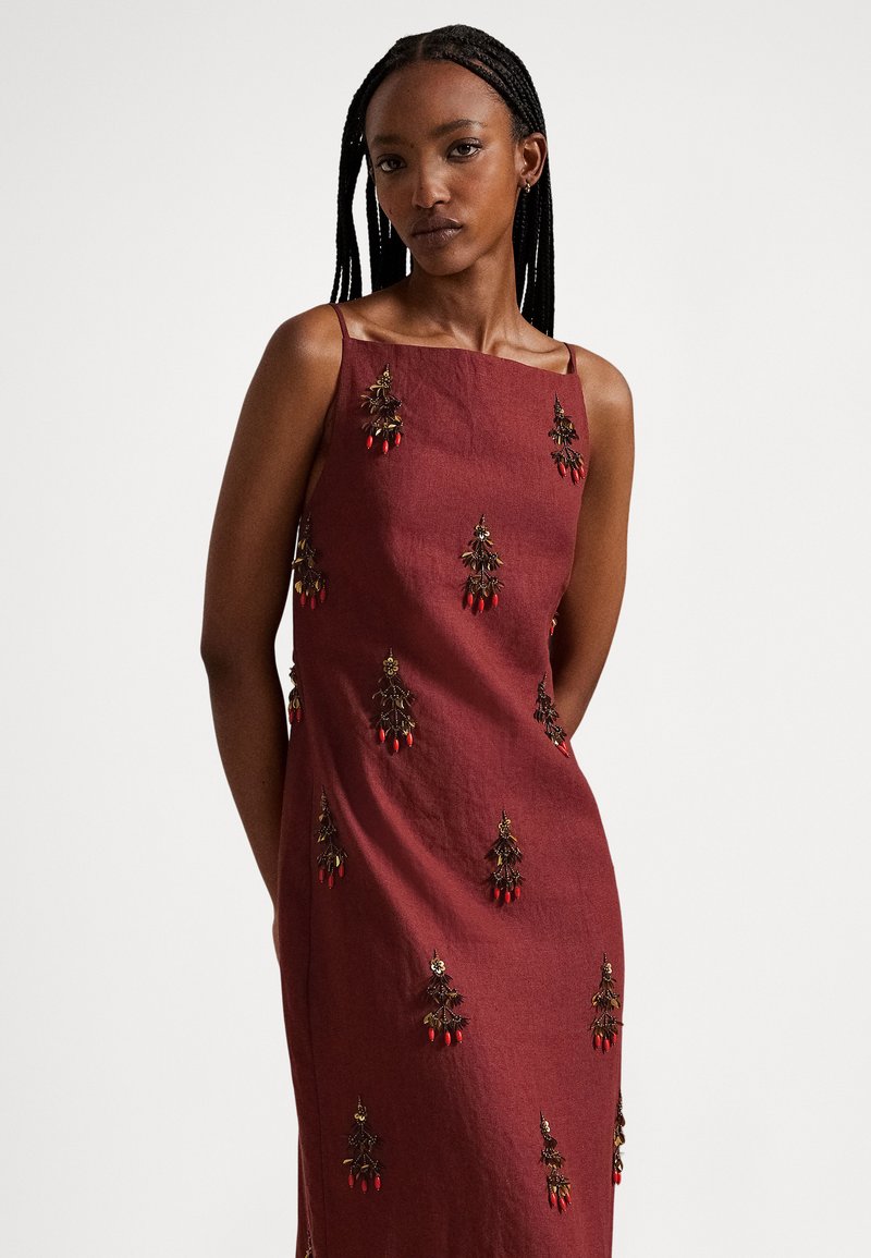 Žena s vrkočmi vo vlasoch, oblečená v bezrukávových bordových šatách zdobených drobnou červenou a zlatou výšivkou, stojaca na jednoduchom pozadí.