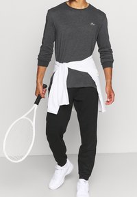 Långärmad mörkgrå skjorta med en grön logotyp, vit jacka knuten i midjan, svarta byxor och vita sportskor, hållande ett tennisracket.