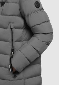 Grå polstret jakke med en glat tekstur, der har en lynlåslomme, et rundt logo-patch påærmet og horisontalt quiltet design.