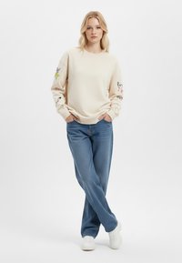 Felpa beige con disegni stampati colorati sulle maniche, abbinata a jeans blu a gamba dritta e sneakers bianche. Stile semplice e casual.