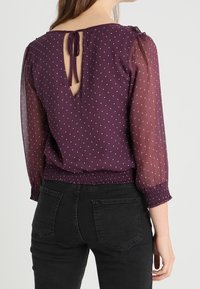 Lila Bluse mit durchsichtigen, gepunkteten Ärmeln, gerafftem Saum und einem Schlüssel-Ausschnitt mit Bindeband. Getragen zu schwarzen Jeans, die eine taillierte Passform zeigen.