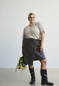 Nais, kes kannab tekstureeritud heledat halli lühikeste varrukatega pluusi, tumedat salli seelikut ja musti põlvikõrguseid saapaid, hoiab käes mitmevärvilist lillekimp.