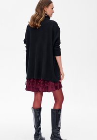 Zwarte oversized gebreide trui, laag over een bordeauxrode rok met ruffles, gecombineerd met zwarte knie-hoge laarzen. Glad textuur en casual pasvorm.
