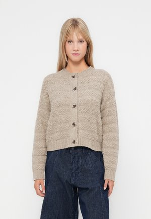 Beige teksturert cardigan med ribbestrikket kant og rund hals, med fem brune knapper foran. Parret med mørkstripete bukser.