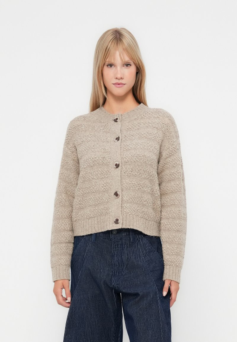 Beige teksturert cardigan med ribbestrikket kant og rund hals, med fem brune knapper foran. Parret med mørkstripete bukser.