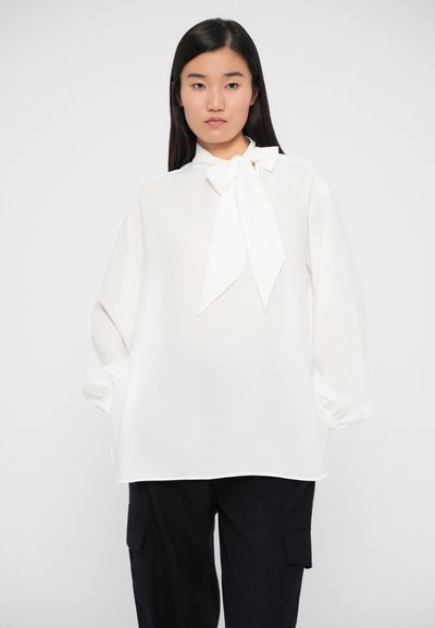 White blouse with a bow tie at the collar, long balloon sleeves, and a loose fit, made from a lightweight fabric. Paired with dark trousers.