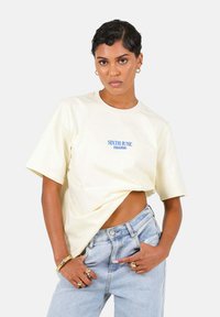Lys gul oversized bomulds t-shirt med blå teksttryk "SIXTH JUNE PARADISO." Sammen med højtaljede lys blå denim jeans.