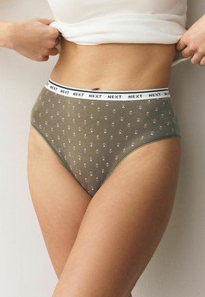 Kobieta w oliwkowo-zielonych figach z wysokim stanem, z małym kwiatowym wzorem i białym paskiem z napisem "NEXT", lekko podnosząca biały top.