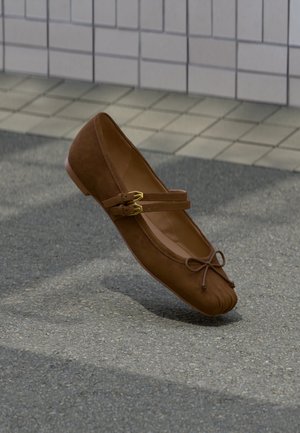 Καφέ μπαλαρίνα με διπλούς ιμάντες και χρυσές αγκράφες, ένα μικρό φιόγκο μπρος, που αιωρείται πάνω από πλακωμένο και ασφαλτοστρωμένο έδαφος.