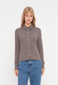 Långärmad, kragskjorta i ribbat, brunt tyg med knappstängning framtill. Matchas med högmidjade blå jeans.