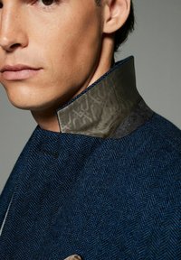 Blaues Fischgrätjackett mit einem strukturierten grauen und gemusterten Kragen. Nahaufnahme der Kragendetails, die die Stoffschichtung und subtile Designelemente zeigen.