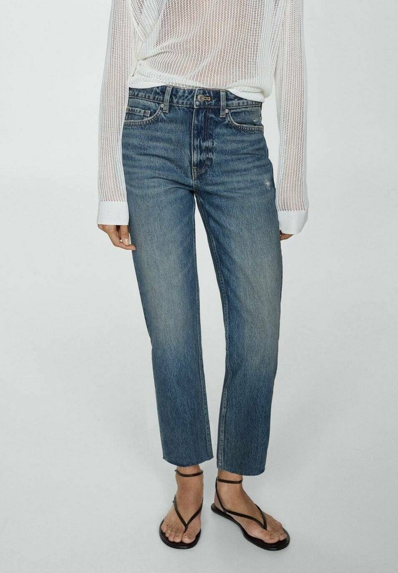 Personne portant un jean bleu taille haute, un haut en tricot blanc manches longues et des sandales plates noires à brides sur un fond uni.