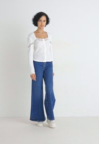 Hvit ribbet knappet cardigan med blonder, parret med blå vide jeans. Modellen har på seg hvite joggesko og står foran en ensfarget bakgrunn.