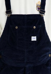 Marineblaues Corduroy-Overall mit verstellbaren Trägern, Metallbeschlägen, Fronttasche und einem weißen "Lee"-Logopatch. Strukturiert sichtbares Material.
