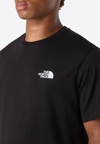 Primer plano de una persona que lleva una camiseta negra con un pequeño logo blanco de "The North Face" en el pecho.