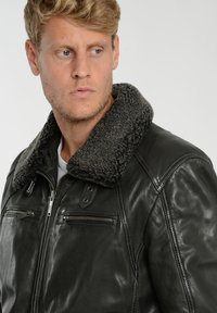 Svart skinnjacka med en tjock, mörkgrå shearlingkrage. Har dragkedjefickor, slät textur och detaljerad sömnad.