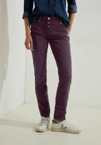 Pantalon en coton violet avec une coupe slim, design à cinq poches et fermeture à cinq boutons, associé à des baskets blanches et une chemise en denim.