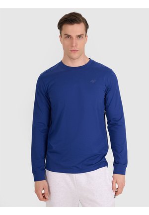 Hombre con camisa azul lisa de manga larga y pantalones de color claro, de pie frente a un fondo liso gris claro.