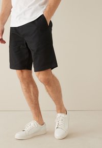 Schwarze Shorts mit geradem Schnitt, ausgestattet mit Seitentaschen und einer glatten Textur, kombiniert mit weißen Sneakern und einem schlichten weißen T-Shirt.