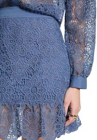Robe en dentelle bleue avec des motifs floraux, des manches longues transparentes et un ourlet à volants. Le tissu présente des détails délicats et une ceinture lisse.